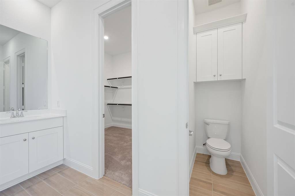 This photo shows a bright, modern primary bathroom with a white vanity and sink, a separate toilet area with overhead storage, and an adjoining walk-in closet with shelving. The room features light wood-look tile flooring.