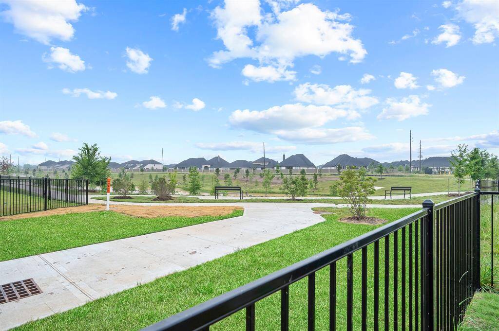 This photo showcases a spacious backyard with a black metal fence, adjacent to a well-maintained pathway and green space. The area is open, with clear views of a suburban neighborhood under a bright blue sky.
