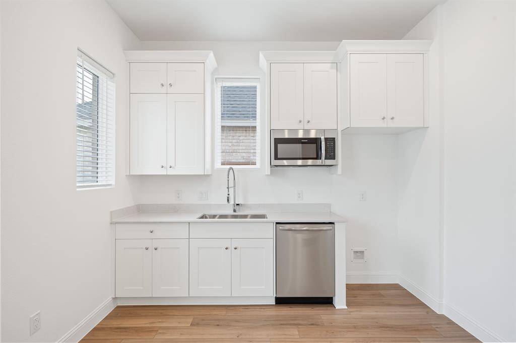 Next Gen® Suite features a modern, compact kitchen with white cabinetry, stainless steel appliances, and wood looked flooring. It includes a sink, a built-in microwave, and a dishwasher, with ample natural light from two windows.