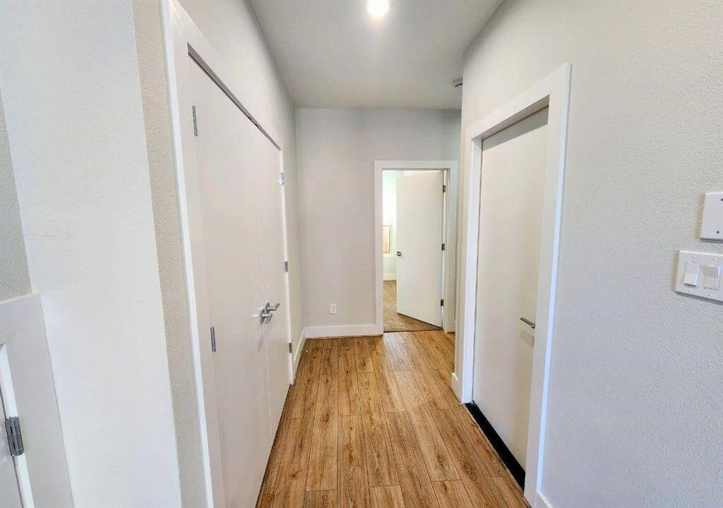 Clean, well-lit hallway with light walls and wood-style flooring. It features two closed doors on either side and an open door at the end leading to another room.