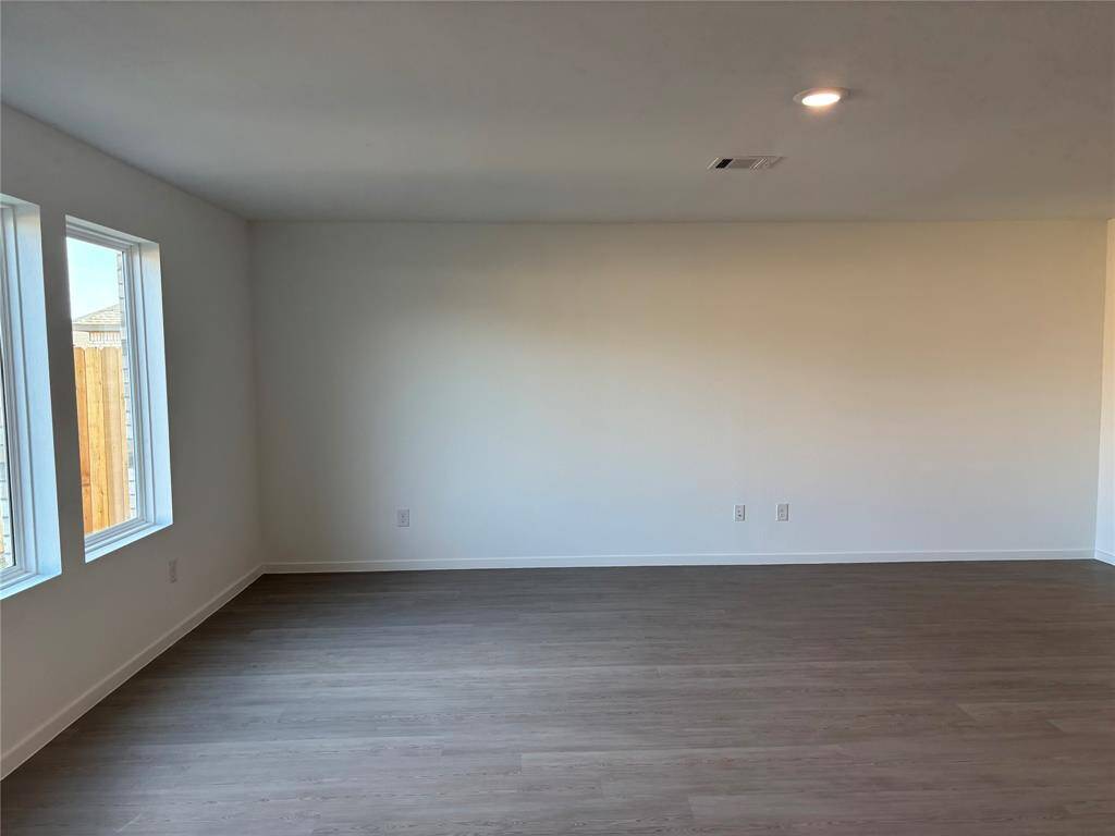 This photo shows a spacious, family room with light-color walls and hardwood flooring. It has large windows allowing natural light and a ceiling light fixture, creating a bright and welcoming space.