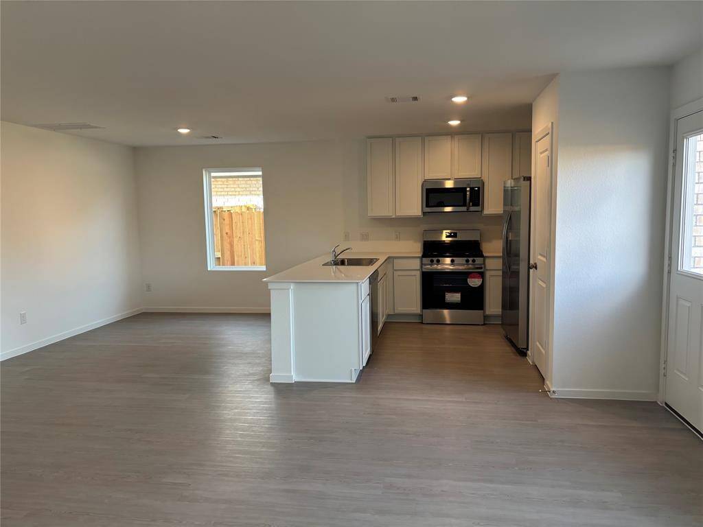 This photo shows a modern open-concept family area with a kitchen featuring stainless steel appliances, white cabinetry, and a center island. The space has light wood flooring and ample natural light from a window, creating a bright and welcoming atmosphere.