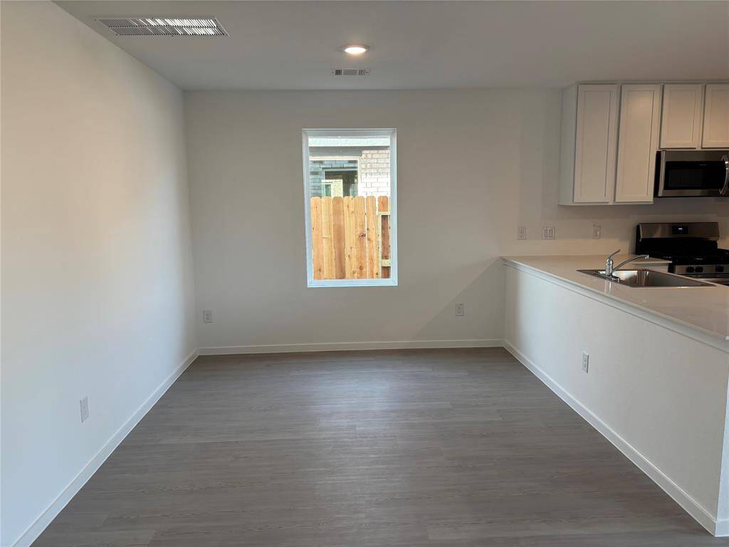This photo shows a bright, open kitchen and dining area with modern appliances, white cabinetry, and light wood flooring. A window provides natural light and a view of the fenced yard.