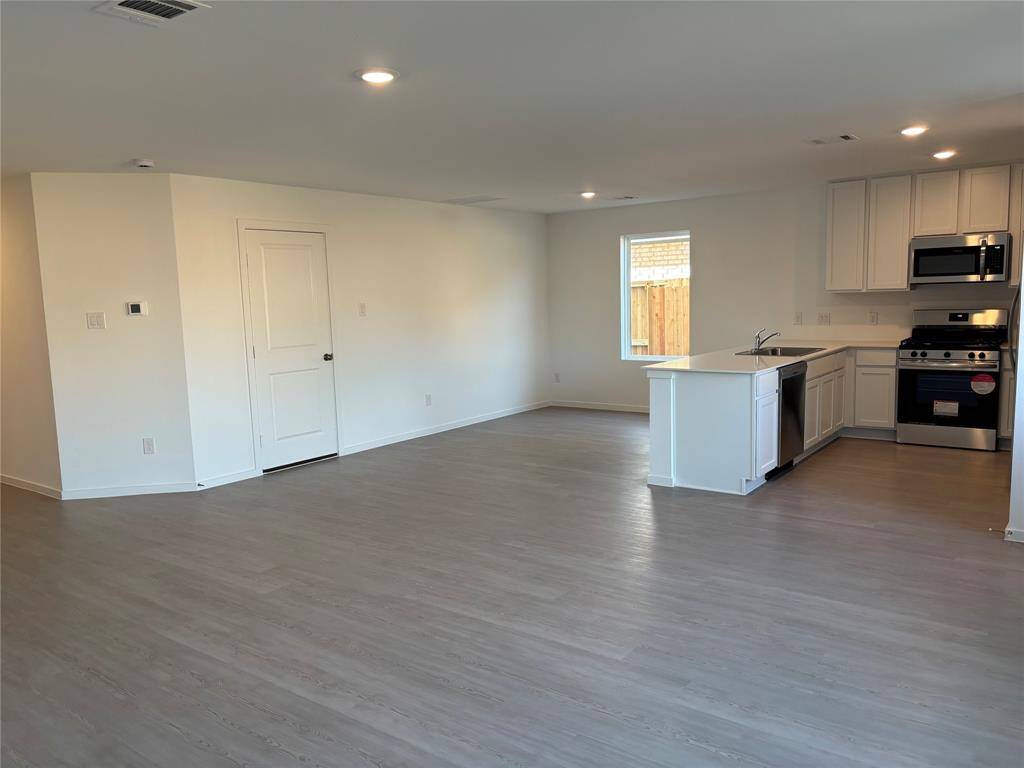This photo shows a modern, open-concept living space with light wood flooring and recessed lighting. The kitchen features white cabinetry, stainless steel appliances, and a central island. A window provides natural light, and the area is spacious for versatile living arrangements.