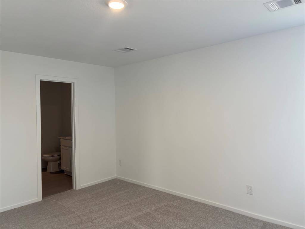 This photo shows a clean, bright primary room with neutral carpeting and a small en-suite bathroom. The walls and ceiling are painted white, creating a fresh and spacious feel.