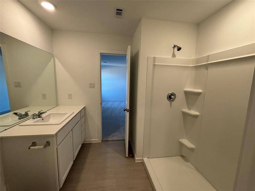 This primary bathroom features a spacious shower, a single-sink vanity with ample counter space and storage, and modern lighting. The design is simple and functional, with a doorway leading to another room.