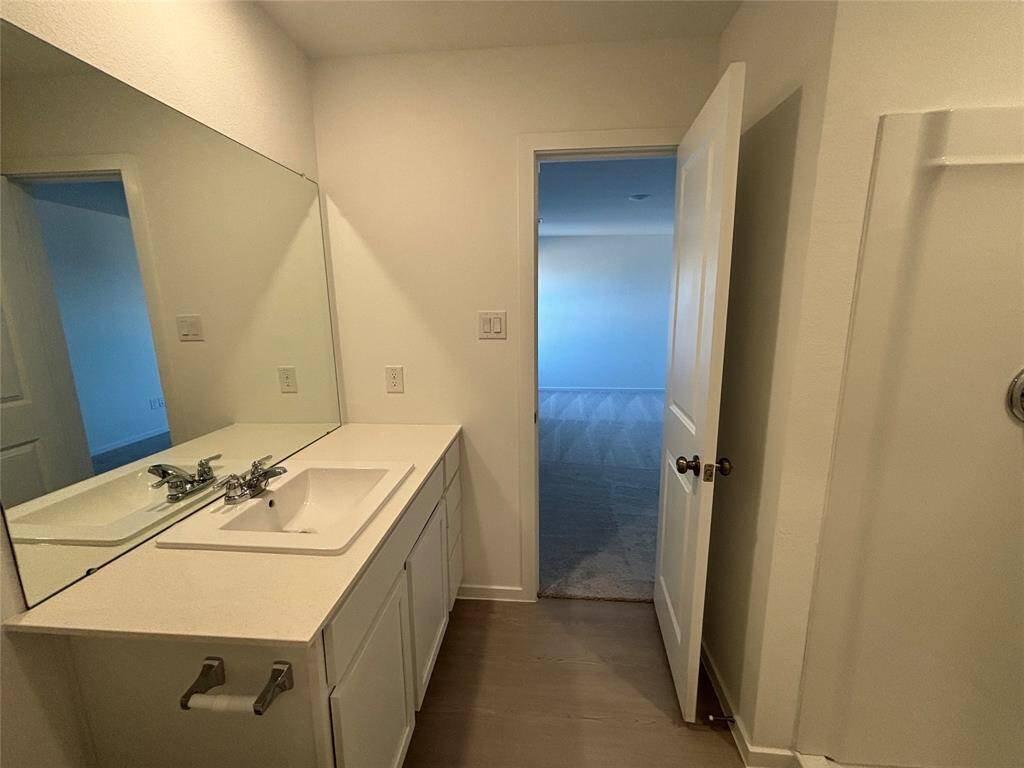 This photo shows a primary bathroom with a large mirror, a single sink vanity with storage, and a door leading to a carpeted room. The space is well-lit and has neutral tones.