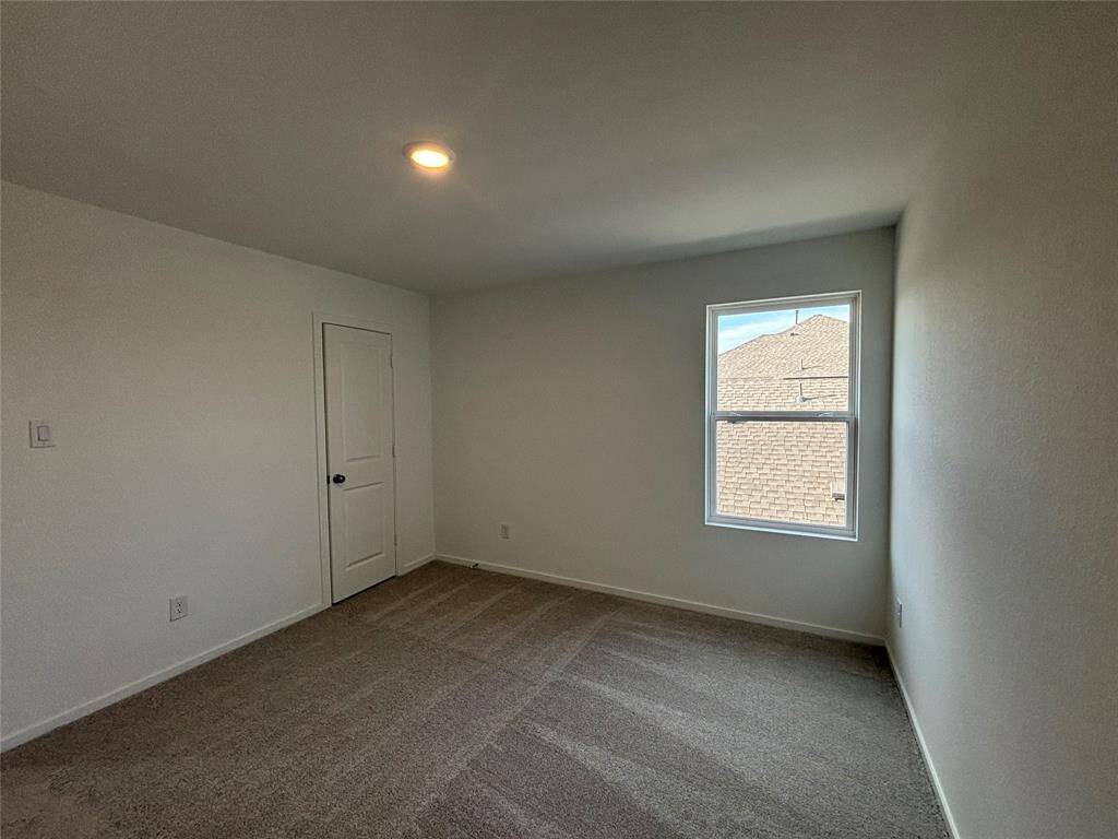 A cozy, well-lit bedroom with neutral walls and carpeting. It features a single window allowing natural light and a door leading to another space.