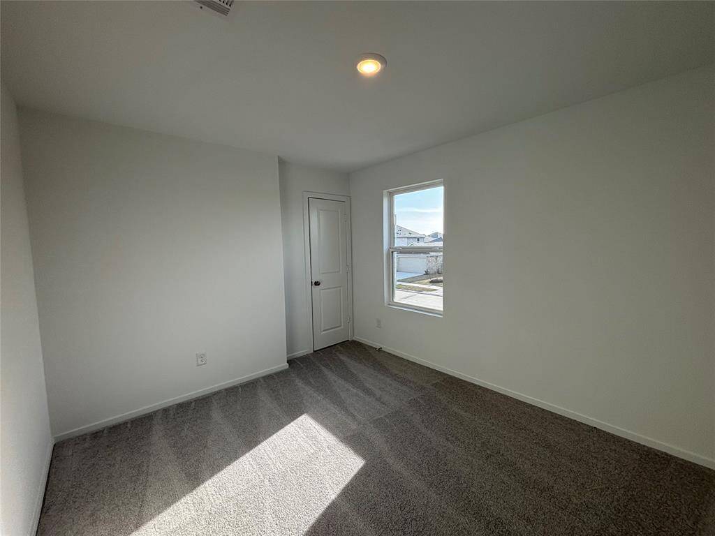 This is a bright, small bedroom with neutral walls and carpet, featuring a window with natural light and a simple door.