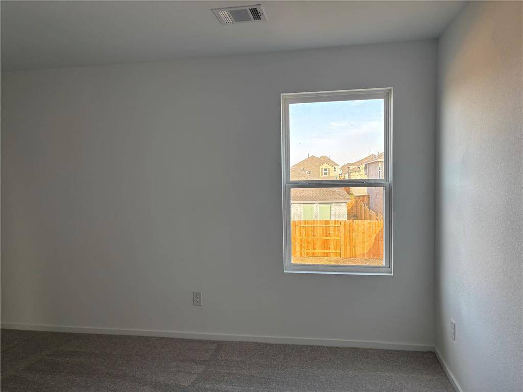 This is a bright, empty room with neutral walls and carpet flooring, featuring a window offering a view of neighboring houses and a wooden fence.