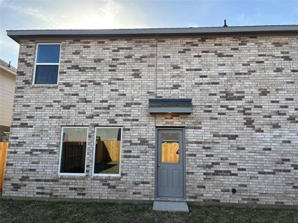 This photo shows the rear exterior of a two-story home with light-colored brickwork. It features a single door and three windows, set within a fenced backyard with a small lawn area.
