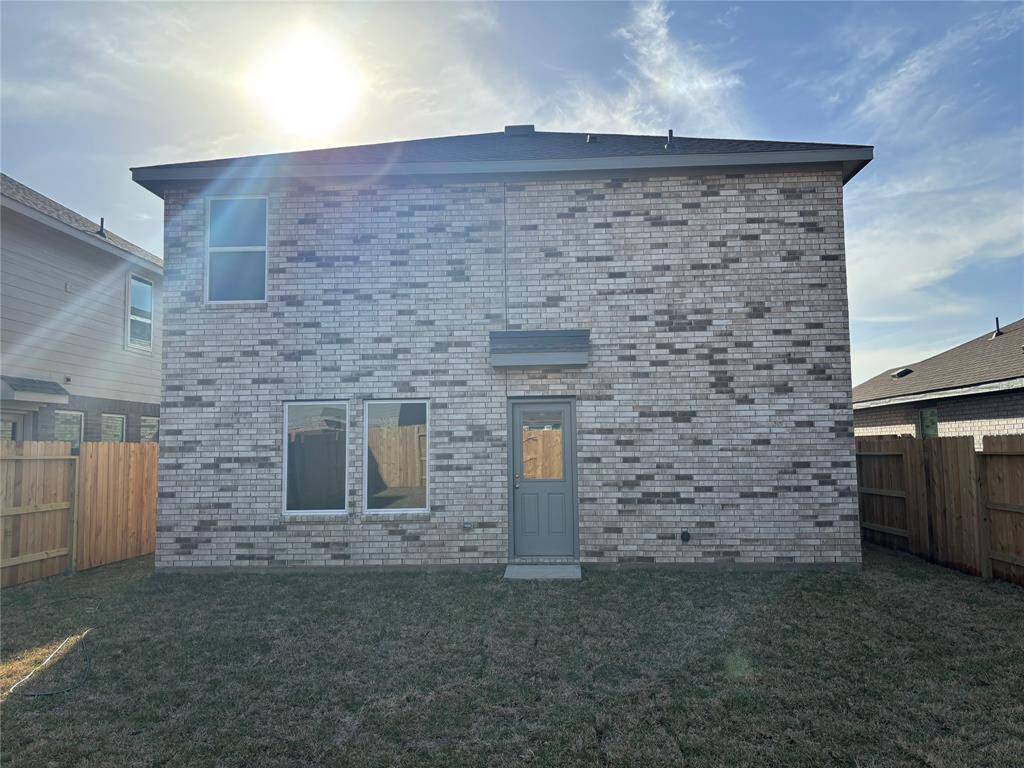 This photo shows the back view of a two-story brick house with a fenced yard. The exterior features a mix of light and dark bricks, two windows, and a central door, all under a clear sky.