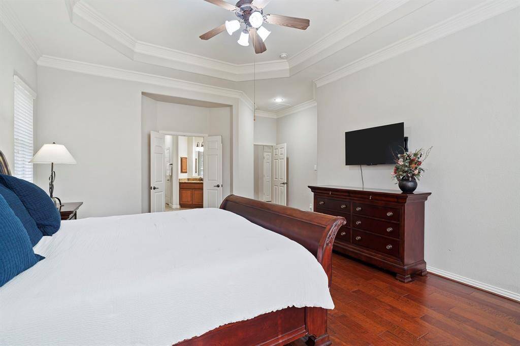 A dramatic tray ceiling, timeless crown molding, and rich wood flooring exude elegance in the primary bedroom. Double doors open to a luxurious en suite bathroom.