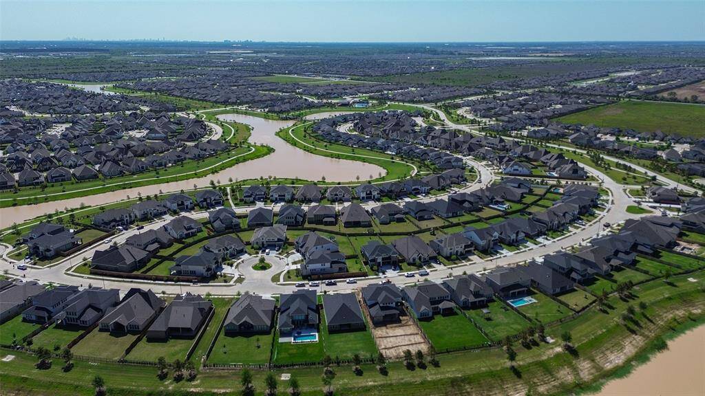 This aerial photo shows a spacious suburban neighborhood with neatly arranged houses, curving roads, and a scenic waterway running through the area. The community features ample green space and some homes with private pools, providing a serene and family-friendly environment.
