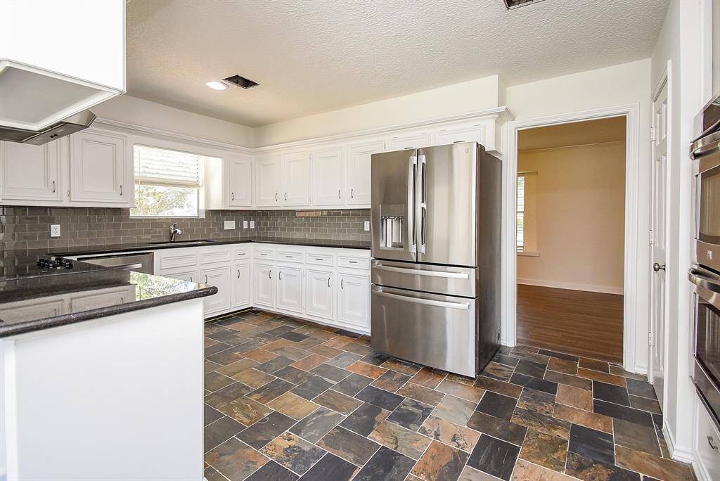 This kitchen features modern stainless steel appliances, white cabinetry, and a dark tile backsplash. The slate tile flooring adds character, while ample natural light comes in through a window above the sink.