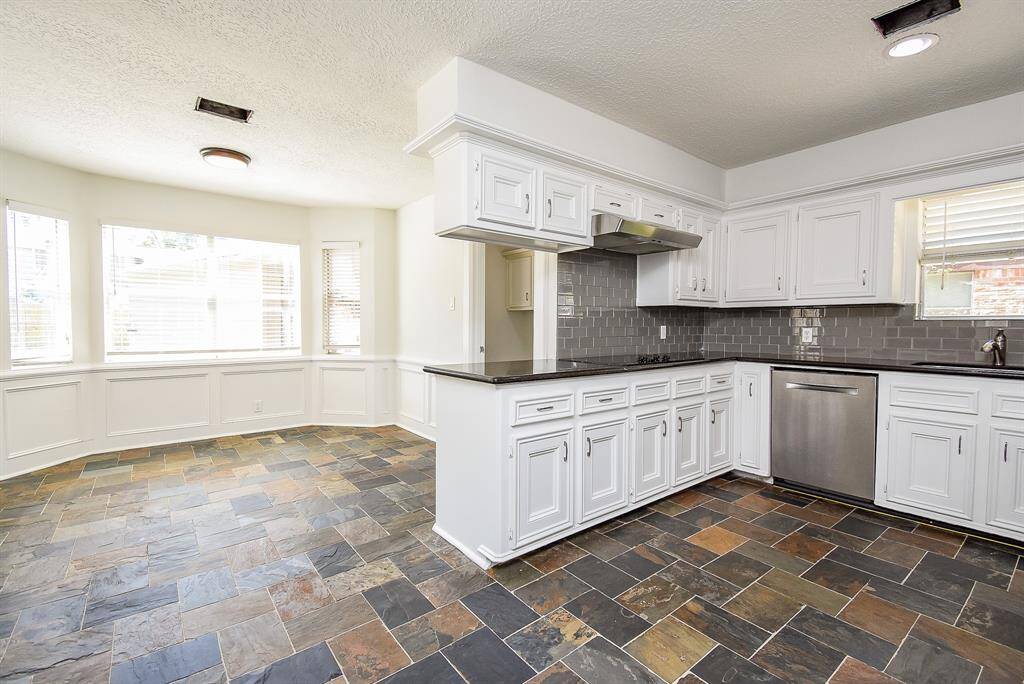 This spacious kitchen features sleek white cabinetry, stainless steel appliances, and a dark tiled backsplash. The open layout offers ample natural light from large windows and a stylish slate floor, perfect for cooking and entertaining.
