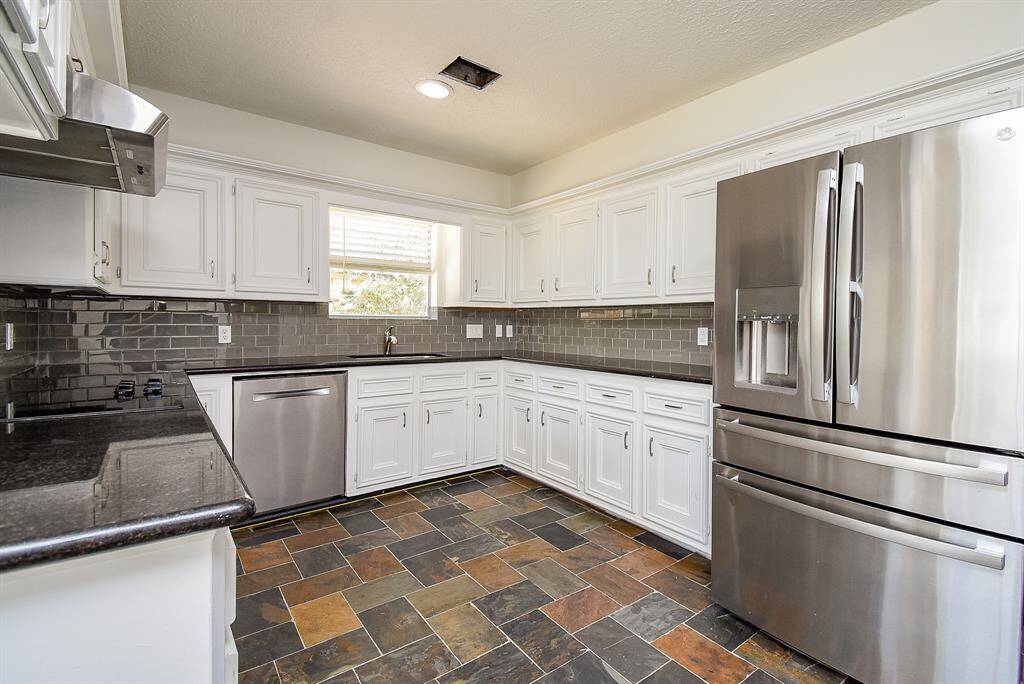 This kitchen features modern stainless steel appliances, white cabinetry, and a sleek dark tile backsplash. The slate tile flooring adds a rustic touch, and the space is well-lit with natural light from the window.