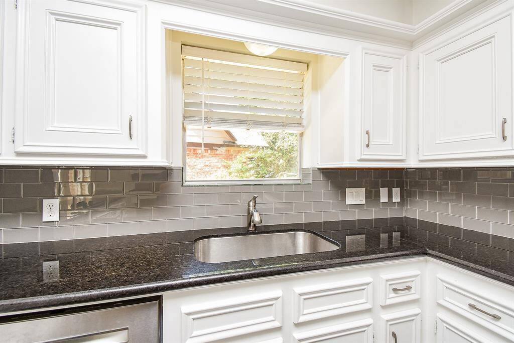 This kitchen features sleek white cabinetry, a modern gray subway tile backsplash, and polished black granite countertops. A stainless steel sink is positioned under a window, providing natural light and a view outside.
