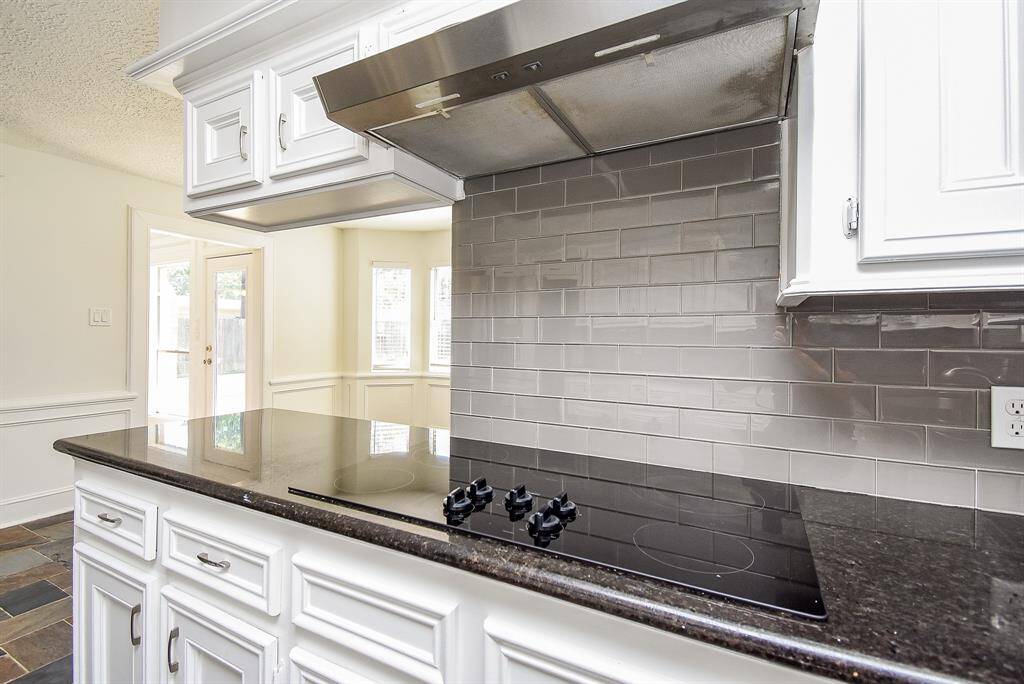Modern kitchen with sleek black countertops, gray subway tile backsplash, and white cabinetry. Features an electric cooktop and stainless steel range hood. Bright and airy with natural light from adjacent windows.