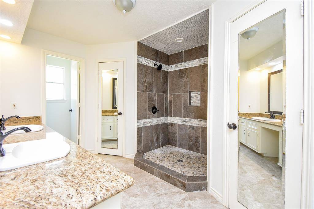 This bright bathroom features a spacious walk-in shower with modern tile design, dual sinks with granite countertops, and a large mirror, creating a luxurious and functional space.