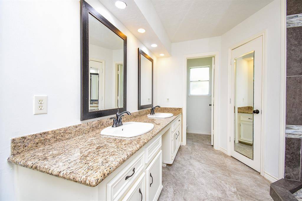 This bathroom features a double sink vanity with granite countertops and framed mirrors. The space is bright with recessed lighting and has a neutral tile floor. A mirrored door adds elegance and functionality.