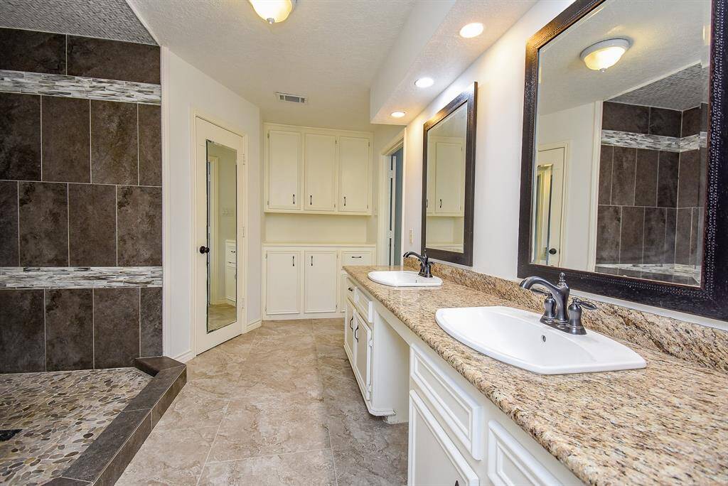 spacious bathroom with dual sinks set in a granite countertop, large mirrors, and ample cabinetry for storage. It features a modern walk-in shower with stylish tile work and pebble flooring.