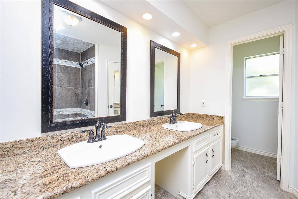 Primary bathroom features a double vanity with granite countertops, framed mirrors, and stylish fixtures. The room has ample lighting and a separate area with a window, offering a bright and functional space.