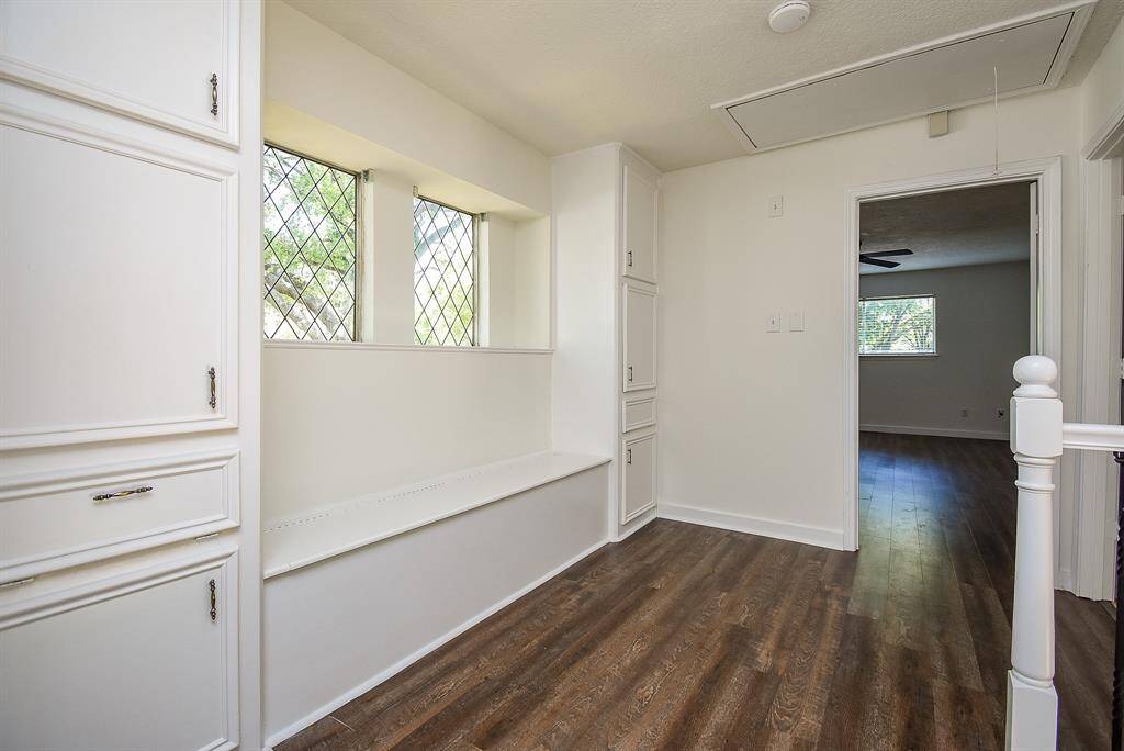 This photo shows a bright hallway with wood flooring and white walls. It features built-in storage cabinets and a charming window seat with diamond-patterned windows. The hallway leads to a room with ample natural light and a ceiling fan.
