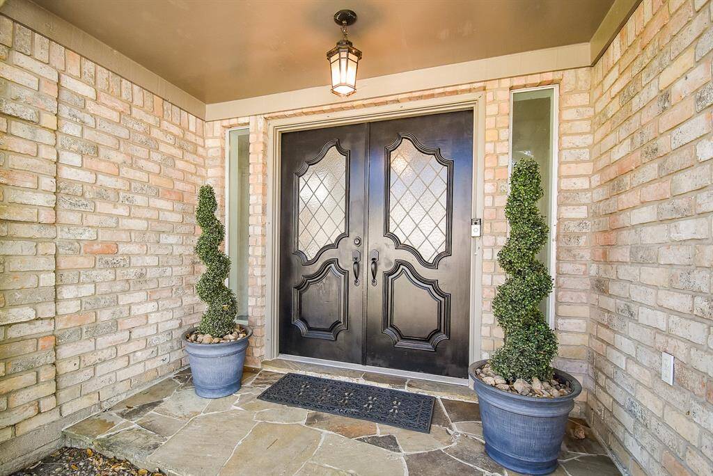 This inviting entryway features a doubles solid wood door with elegant glass panels that allow natural light to pour in the entry way. The brick exterior and stone flooring add a touch of charm, complemented by a warm overhead light.