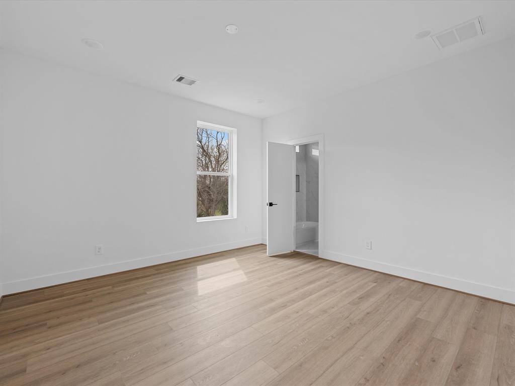 This photo shows a bright, spacious room with light wood flooring, white walls, and a window providing natural light. There is an open door leading to a bathroom with modern finishes.