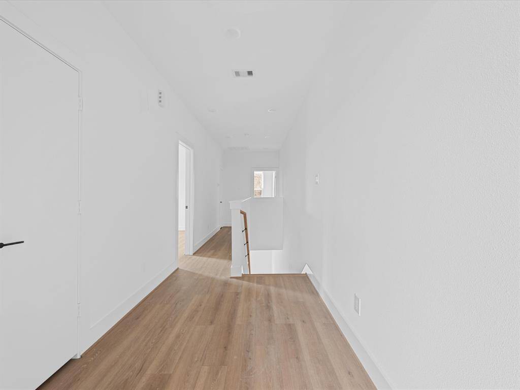 Bright, modern hallway with light wood flooring, white walls, and a staircase leading down. Doorways on the left suggest access to additional rooms.