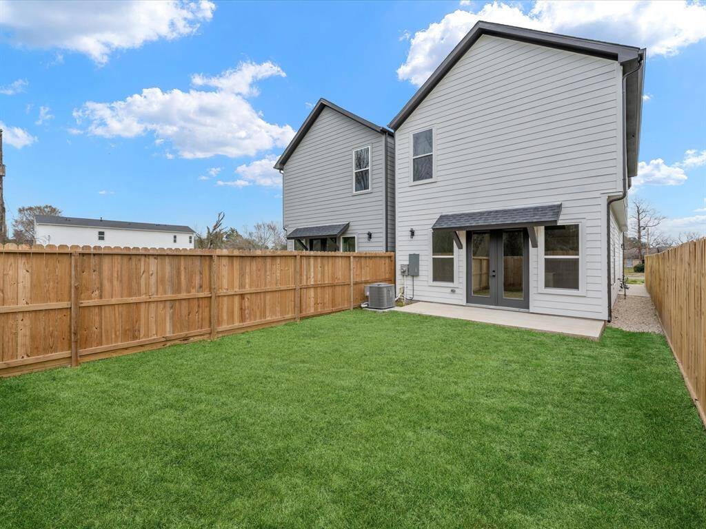 This photo shows a backyard with a well-maintained lawn, a wooden privacy fence, and an outdoor HVAC unit. There is a concrete patio area adjacent to the house, offering space for outdoor activities. The setting is open and bordered by trees, providing a private and serene atmosphere.