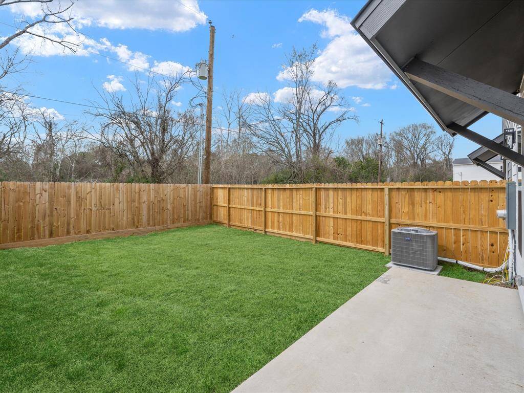 This bright living room features a modern design with light wood flooring, a large sectional sofa, and a cozy ottoman. It has a wall-mounted TV, stylish decor, and large windows that let in natural light, leading to a fenced backyard through elegant French doors.