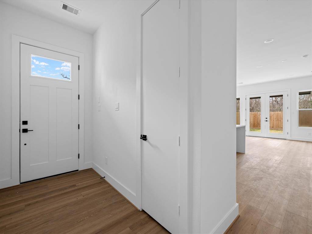 A bright, minimalist hallway with light wood flooring and white walls. There are multiple doors, including a dark-colored one with a modern handle, adding a sleek touch to the space.