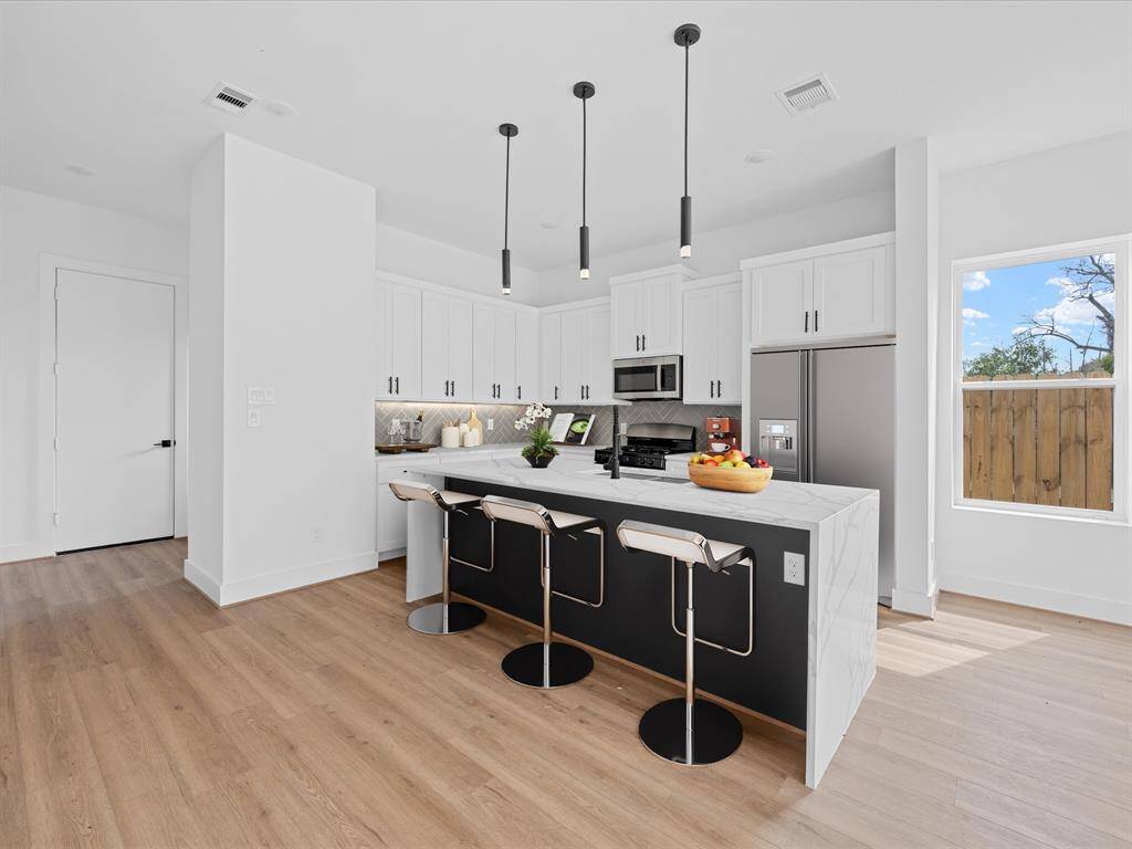 This photo showcases a spacious, modern open-concept living area with light wood flooring and abundant natural light. The kitchen features a large island, sleek white cabinetry, and stainless steel appliances. A staircase leads to the upper level, enhancing the contemporary design.