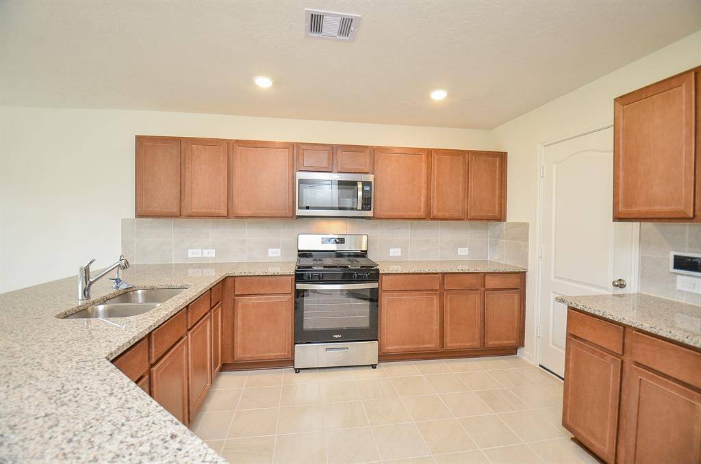 This kitchen features a spacious layout with ample granite countertops and wooden cabinetry. It includes stainless steel appliances, a double sink, and tiled flooring, offering a modern and functional cooking space.