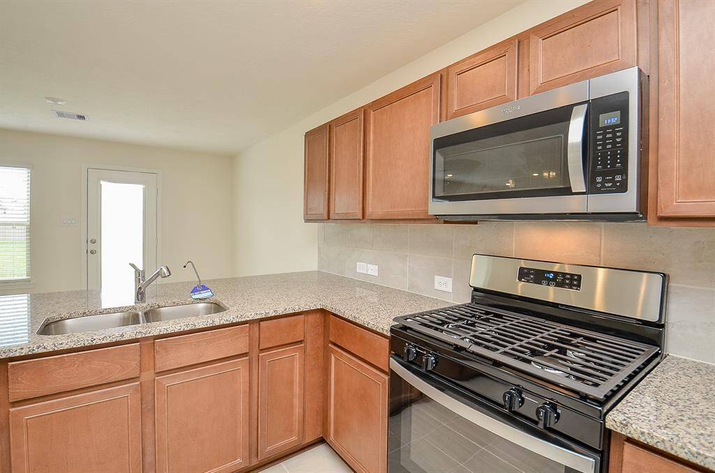 This kitchen features granite countertops, stainless steel appliances, and ample wooden cabinetry. It includes a double sink, a gas stove, and a built-in microwave, with a door leading to the backyard, offering plenty of natural light.