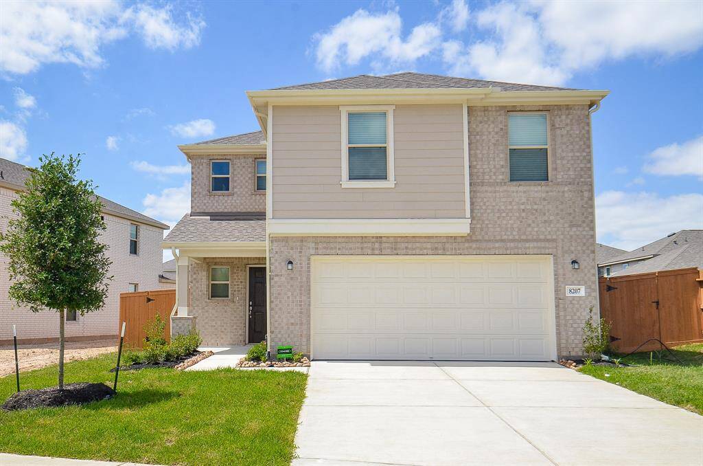 This is a modern two-story home with a brick exterior, featuring a spacious two-car garage. The front yard is landscaped with a small tree and shrubs, and the property is enclosed with a wooden fence. The house has a clean, inviting appearance under a bright blue sky.
