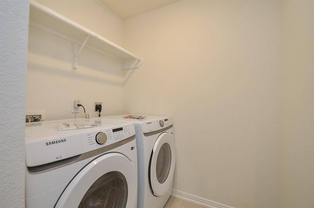 This photo shows a compact laundry room with a Samsung washer and dryer set, a light beige wall, and a white shelf above for storage.