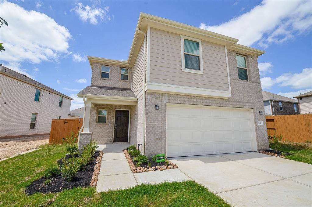 This photo shows a modern two-story house with a light brick and siding exterior. It features a double garage, a small front porch, and a neatly landscaped front yard. The home is situated in a suburban neighborhood.