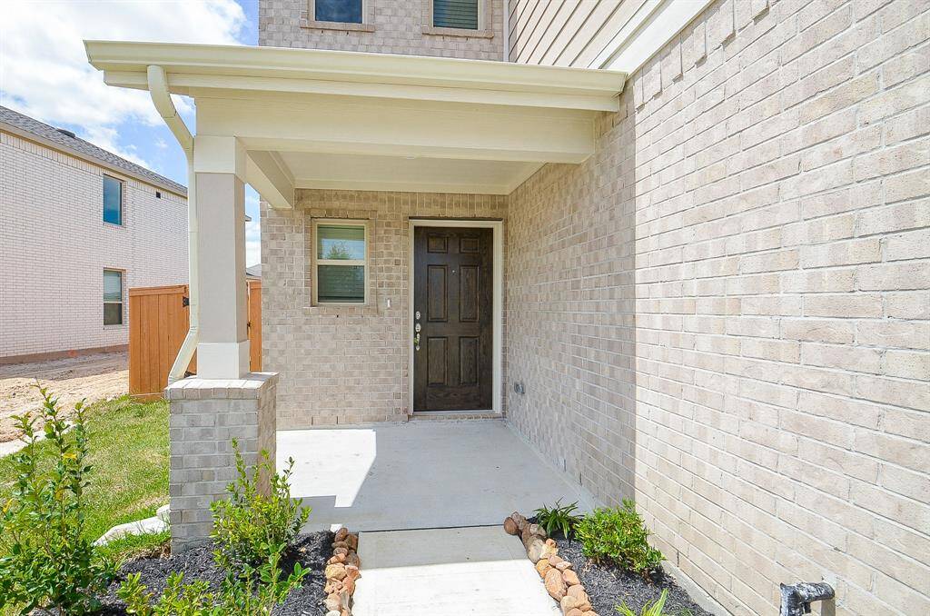 This photo shows a welcoming front porch of a modern brick home, featuring a solid wooden door, a small window, and simple landscaping with shrubs. The covered entryway provides shelter and adds to the home's curb appeal.