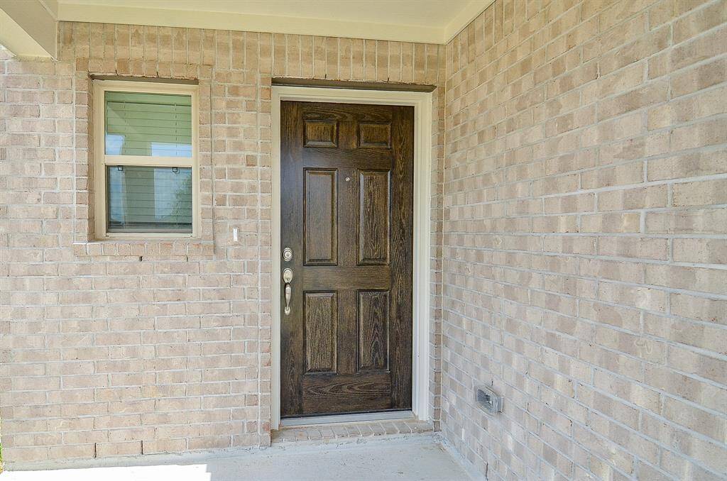 This photo shows a welcoming front entry featuring a sturdy wooden door with a classic design, set against a neutral brick exterior. A small window beside the door adds natural light to the entryway.
