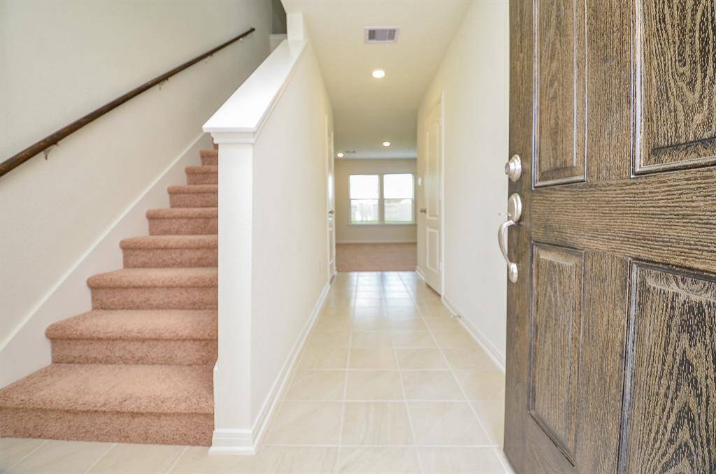 This photo shows a welcoming entrance with a wooden door opening into a bright hallway. To the left, there's a carpeted staircase leading upstairs, and the hallway features tiled flooring, leading to a well-lit room with large windows.