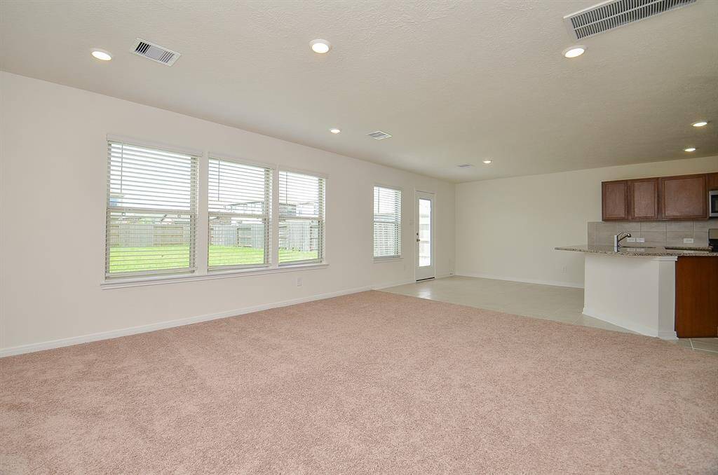 Spacious, open-concept living area with large windows offering plenty of natural light. Features a combination of carpeted and tiled flooring, leading to a modern kitchen with granite countertops and wooden cabinetry. Ideal for comfortable living and entertaining.