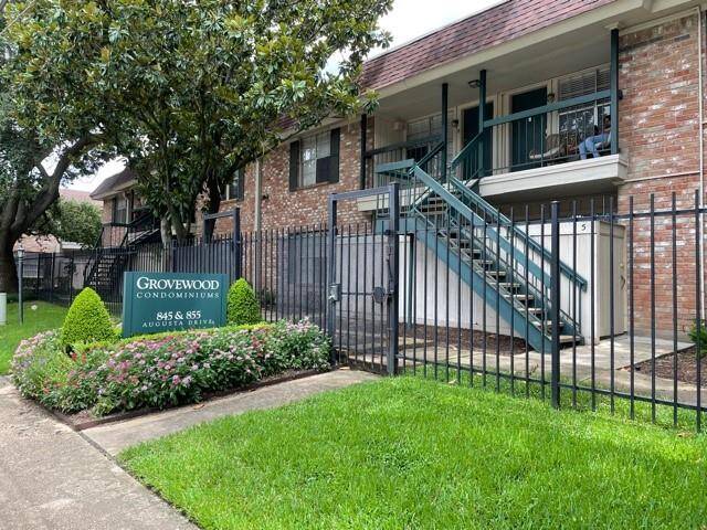 Grovewood Condominiums, showcasing a well-maintained brick building with exterior stairs and a landscaped front area. The green lawn and flowering plants add to the curb appeal, while a secure black metal fence borders the property.
