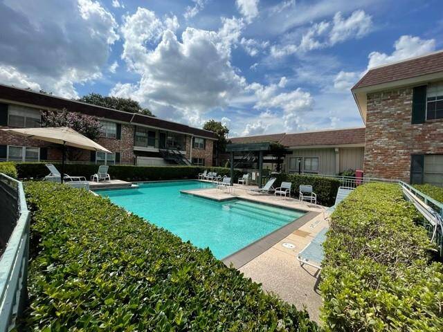 Well-maintained outdoor pool area in a residential complex. Surrounded by lush greenery and brick buildings, the space is equipped with lounge chairs and umbrellas, offering a relaxing environment. Perfect for leisure and entertaining, with clear blue skies enhancing the inviting atmosphere.