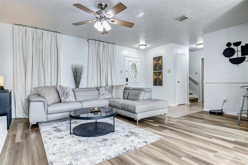 This inviting living room features modern decor with a neutral color palette, a cozy sectional sofa, and a stylish round coffee table. The flooring adds warmth, complemented by a contemporary ceiling fan. The space is well-lit with both natural and ambient lighting, creating a welcoming atmosphere.