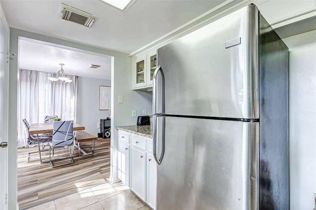 This photo showcases a bright kitchen area with a stainless steel refrigerator, granite countertops, and white cabinetry. It opens into a dining space with modern chairs, a wooden table, and natural light streaming through large windows with sheer curtains.