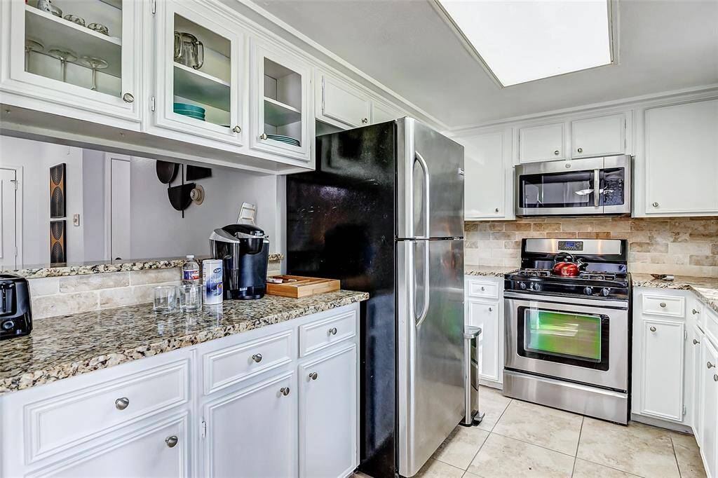 This kitchen features white cabinetry with glass-front uppers, granite countertops, and stainless steel appliances, including a fridge and gas stove. The backsplash is a neutral stone tile, and the flooring is light-colored tile, creating a bright and modern space.