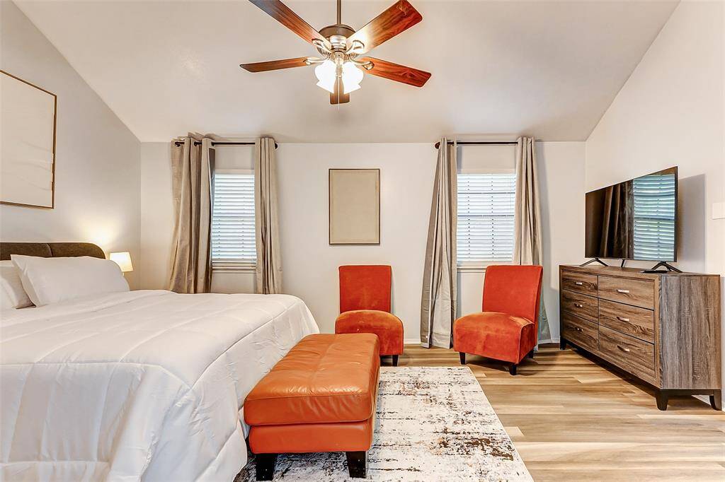This cozy bedroom features a comfortable bed, two bright orange chairs, and a matching ottoman. It has a ceiling fan, ample natural light from two windows with curtains, a large dresser, and a mounted TV. The room offers a warm, inviting atmosphere with neutral tones and  flooring.